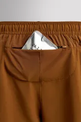 Pantalón corto deportivo color marrón oliva, confeccionado en tejido técnico ligero y elástico, con forro interior. Presenta cintura elástica ajustable con cordón, bolsillos frontales, bolsillo de seguridad trasero con cierre y aberturas laterales en el bajo.