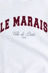 Buzo gris melange con cuello redondo, hombros caídos y puños y bajo acanalados. Estampado frontal en color bordó con el texto "LE MARAIS" y "Ville de Paris 1995".