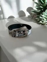 Cinto con estampado de piel de serpiente en blanco y negro, con hebilla metálica grande de estilo western con detalles grabados.