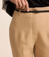 Pantalón de sastrería corte carrot, color beige, con cintura alta, pinzas frontales y cinturón fino incorporado con hebilla metálica.