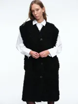 Chaleco largo reversible de diseño versátil. Por un lado, presenta una textura de peluche sintético en color marrón, y por el otro, un acabado liso. Cuenta con cierre frontal de botones y corte recto.