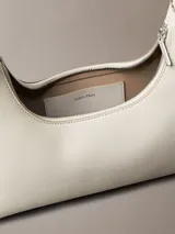 Cartera de hombro Calvin Klein de diseño estructurado en color beige, con cierre de cremallera superior, correa ajustable y detalle del logo de la marca grabado en la parte frontal.