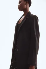 Blazer largo negro, de corte recto y botonadura sencilla con dos botones frontales. Presenta solapas de muesca, bolsillos delanteros ribeteados con solapa y hombreras.