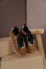 Championes urbanos de gamuza color marrón, con detalles en charol negro y bordes dorados. Presentan cordones negros y suela de goma al tono.