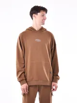 Hoodie color marrón con capucha, bolsillo canguro frontal y puños y cintura acanalados. Presenta un pequeño parche bordado con texto en el pecho.