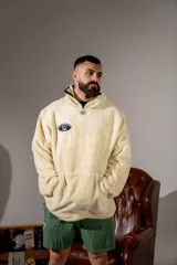 Canguro de peluche color crema, con cuello alto y cierre frontal parcial. Presenta un parche ovalado con diseño gráfico en el pecho y un bolsillo frontal tipo canguro.