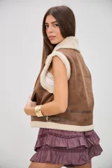 Chaleco de estilo aviador en color camel, confeccionado en material efecto cuero con interior y bordes de corderito sintético color crema. Presenta cierre frontal, cuello solapa y detalles de hebillas metálicas en los laterales y el cuello.