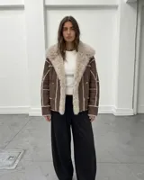 Campera tipo gamulán de cuero marrón con interior y solapas de piel sintética en color crema. Presenta un diseño de paneles con costuras visibles y cierre frontal.