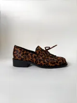 Mocasín de cuero marrón con textura granulada, diseño de horma cuadrada, detalle de flecos y lazo decorativo en el empeine, montado sobre una suela y taco bajo de color negro.