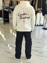 Canguro oversize de color beige claro con capucha, mangas largas y puños elastizados. Presenta un diseño gráfico en la espalda con el texto 'Creative Thinking' en tipografía cursiva negra sobre un fondo de letras grandes en tono rosado.