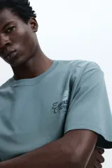 Remera de algodón verde militar, de corte holgado, con cuello redondo y mangas cortas. Cuenta con un pequeño estampado gráfico con texto en el pecho.