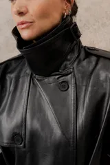 Trench largo de ecocuero negro, con cuello clásico, cierre cruzado con botones laterales y cinturón ajustable.