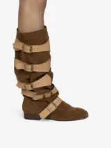 Bota de montar de caña alta y diseño holgado, confeccionada en cuero con acabado de gamuza color marrón. Presenta múltiples correas de cuero liso en color beige con hebillas metálicas doradas a lo largo de la caña y el empeine. Posee un taco bajo de aproximadamente 2,5 cm.