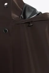 Chubasquero largo color marrón oscuro, con cuello subido y capucha ajustable con cordones. Tiene cierre frontal con cremallera oculta y botones a presión, y bolsillos delanteros con solapa y botón a presión oculto.