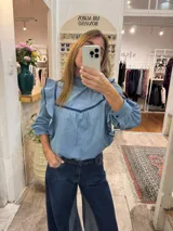 Blusa de jean fino color celeste con detalles de ribetes en terciopelo azul oscuro, cuello alto con volados y mangas largas con volados en los hombros.