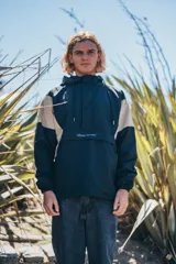 Buzo rompevientos con capucha, diseño tipo canguro con cierre frontal parcial y bolsillo central. Presenta un diseño bicolor en negro y beige, con puños elásticos y cordones ajustables en la capucha.