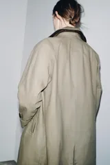 Trench coat de corte largo y diseño minimalista, confeccionado en tejido encerado color beige. Presenta cuello solapa, cierre frontal con botones oscuros y bolsillos laterales integrados.