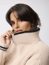 Sweater de tejido grueso en color beige, con cuello alto y cierre frontal parcial. Presenta detalles de rayas negras en contraste en el borde del cuello y en los puños.