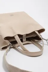 Bolso de mano grande tipo tote de Bimba y Lola, color beige, con doble asa superior y correa ajustable de tejido. Presenta logo metálico en la parte inferior y cierre superior.