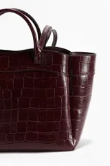 Bolso tipo shopper de cuero sintético con textura de cocodrilo en tono borgoña oscuro. Presenta dos asas de mano cortas y dos asas de hombro largas. La parte superior tiene un borde ondulado.