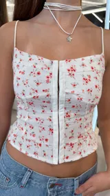 Top blanco de tirantes finos con estampado de pequeñas flores rojas, escote recto y cierre frontal de ganchos con detalle de ribete ondulado en el borde inferior.