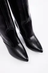 Bota de caña alta confeccionada en cuero vegano color negro, con punta fina y taco cónico de altura media.