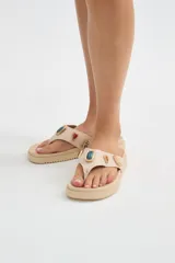 Ojotas beige de plataforma plana con tira de dedo ancha decorada con apliques de piedras de colores en tonos verde, rojo y dorado con bordes metálicos.