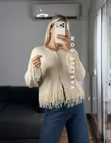 Cárdigan de tejido de punto color crema, con cuello redondo, mangas largas y cierre frontal con botones metálicos. Presenta detalles de flecos en los puños y en el ruedo.