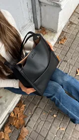 Cartera de mano tipo tote en color negro, confeccionada en cuero con textura granulada. Presenta un diseño minimalista con doble asa superior y un compartimento interior en tono contrastante.