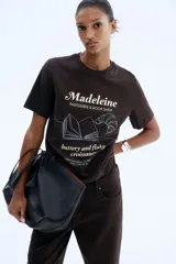 Remera marrón de corte recto con cuello redondo y mangas cortas. Estampado frontal con el texto "Madeleine PASTISSERIE & BOOK SHOP", el dibujo de un libro abierto, un croissant y la frase "buttery and flaky croissants".
