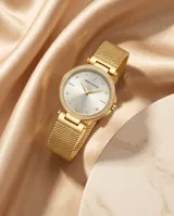 Reloj analógico de mujer con caja redonda dorada, bisel con incrustaciones de cristales y malla de acero tipo milanesa. Esfera minimalista con índices puntuales.