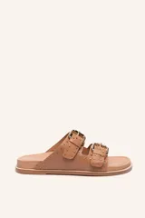 Sandalias planas color tostado con dos tiras anchas ajustables con hebillas doradas y detalles de tachas doradas.