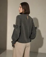 Blusa de color verde oscuro con cuello alto con volados, detalle de lazos frontales y mangas largas abullonadas con lazos decorativos en los codos y puños ajustados.