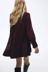Vestido corto de tejido plisado color bordó, con cuello redondo y cierre frontal con botones. Presenta mangas largas tipo globo con puños ajustados y diseño holgado.
