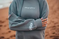 Hoodie gris con capucha y cordones ajustables, de corte holgado y manga larga. Presenta un bordado de flores blancas en la espalda y en el puño.