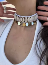 Gargantilla choker con hilo trenzado color blanco, adornada con conchas marinas tipo buzios y medallitas bañadas en oro y acero quirúrgico. Se cierra con cadena de acero quirúrgico y ganchito para ajustar el largo.