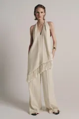 Top halter color crudo, de escote en V y diseño confeccionado en hilatura con mezcla de viscosa. Presenta bajo asimétrico con flecos al tono y cierre en la espalda.