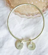 Pulsera rígida dorada tipo esclava, elaborada en bronce, con dos dijes circulares colgantes.