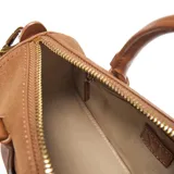 Cartera tipo baúl compacta de símil gamuza color tostado, combinada con detalles en PU liso del mismo tono. Cuenta con doble asa corta rígida, correa larga regulable y desmontable, y herrajes metálicos dorados. El compartimento principal tiene cierre superior y un bolsillo interno con cierre. Presenta el logo XL metalizado en la parte frontal.