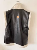 Chaleco negro de cuerina con interior de corderito sintético, bolsillos frontales y hebillas decorativas.