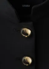 Chaqueta corta negra de cuello alto mao, con cierre frontal cruzado de botones dorados metálicos, hombreras estructuradas y mangas largas con botones en los puños.