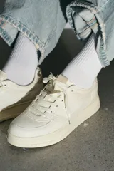 Zapatillas deportivas de mujer, color blanco hueso, con detalles en tono beige claro en el talón y paneles laterales. Presentan una suela de plataforma gruesa y cierre con cordones.