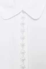 Camisa de popelín color blanco con cuello solapa redondeado y manga larga. Presenta un cierre frontal con botones ocultos y una tapeta decorada con perlas.