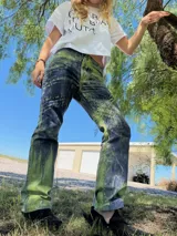 Pantalón de tiro medio y corte recto, confeccionado con un proceso manual que genera un estampado único con variaciones de color en tonos verde y azul oscuro, con detalles de escritura manuscrita en la pierna.