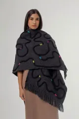 Ruana de tejido de punto grueso en color gris oscuro, con diseño de líneas curvas en relieve de color negro y detalles de costuras decorativas en colores vibrantes. Presenta terminaciones con flecos en el ruedo y una etiqueta de cuero sintético en la parte inferior.