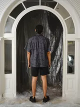 Camisa de manga corta color gris oscuro con estampa exclusiva de símbolos, animales y elementos de la marca en tono negro. Presenta cuello camisero, dos bolsillos delanteros con solapa, corte relajado y cierre frontal con botones metálicos a presión.
