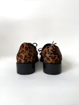 Mocasín de cuero marrón con textura granulada, diseño de horma cuadrada, detalle de flecos y lazo decorativo en el empeine, montado sobre una suela y taco bajo de color negro.