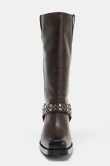 Bota de caña alta en piel color marrón oscuro, con detalle de tira con tachas y abalorios alrededor del tobillo, acabado en punta cuadrada y tacón plano.