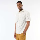 Camisa de manga corta color blanco con diseño estampado de pequeñas palmeras negras, cuello camisero y cierre frontal de botones.