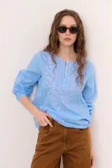 Blusa celeste de manga larga con delicados bordados florales en tono más claro en la zona del pecho y mangas. Presenta escote en V y puños elásticos abotonados.